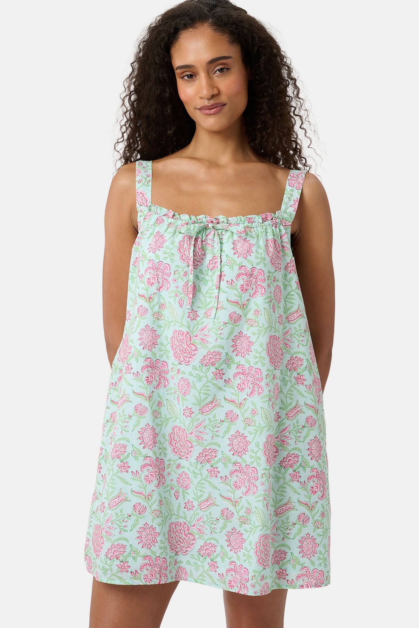 A woman with curly hair wears the Camellia Block Keaton Dress by Roller Rabbit, a sleeveless, loose-fit 100 cotton sundress with pink floral print on a light green background. She smiles softly with her hands behind her back.