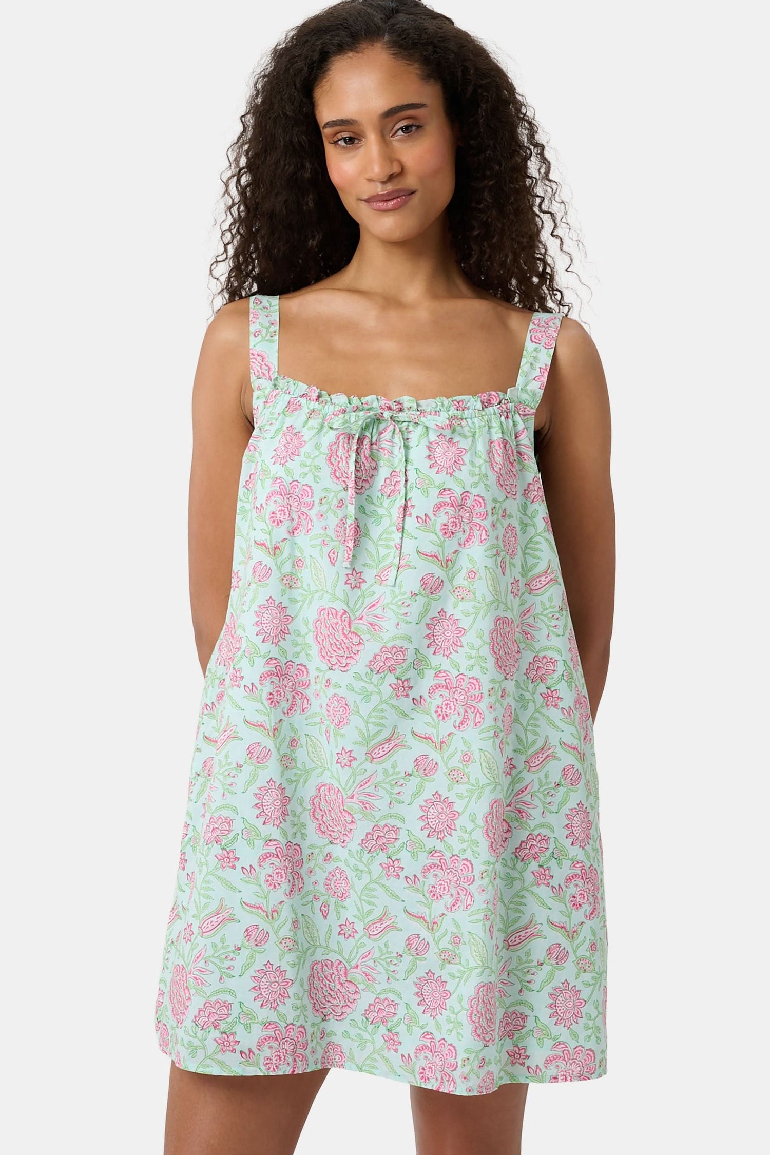 A woman with curly hair wears the Camellia Block Keaton Dress by Roller Rabbit, a sleeveless, loose-fit 100 cotton sundress with pink floral print on a light green background. She smiles softly with her hands behind her back.