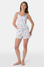A woman with long brown hair wears the Roller Rabbit Camp RR Mischa Set, a light-hued Pima cotton pajama set with playful prints. The adjustable strap tank and shorts provide comfort as she stands turned slightly to the side, gazing off-camera.