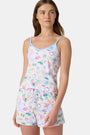 A woman with long brown hair wears the Roller Rabbit Camp RR Mischa Set, a light-hued Pima cotton pajama set with playful prints. The adjustable strap tank and shorts provide comfort as she stands turned slightly to the side, gazing off-camera.