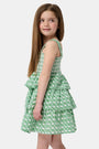 A young girl with curly hair wears the Roller Rabbit Kids Cane Kenia Dress, a green and white patterned cotton tiered dress with wide straps, standing against a plain light background.