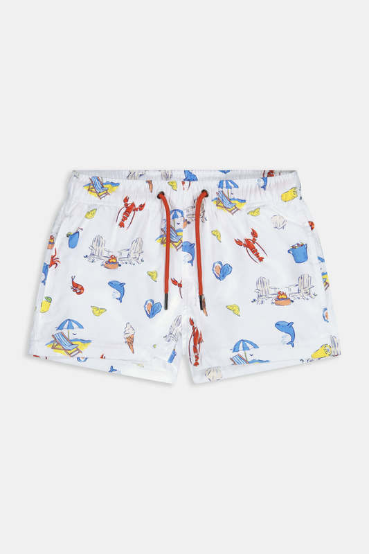 A young boy smiles barefoot in front of a cream-colored vehicle, wearing Roller Rabbit Boys Catch of the Day Shortie Swim Trunk with colorful prints, while another child sits partially visible beside him.