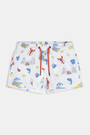 A young boy smiles barefoot in front of a cream-colored vehicle, wearing Roller Rabbit Boys Catch of the Day Shortie Swim Trunk with colorful prints, while another child sits partially visible beside him.