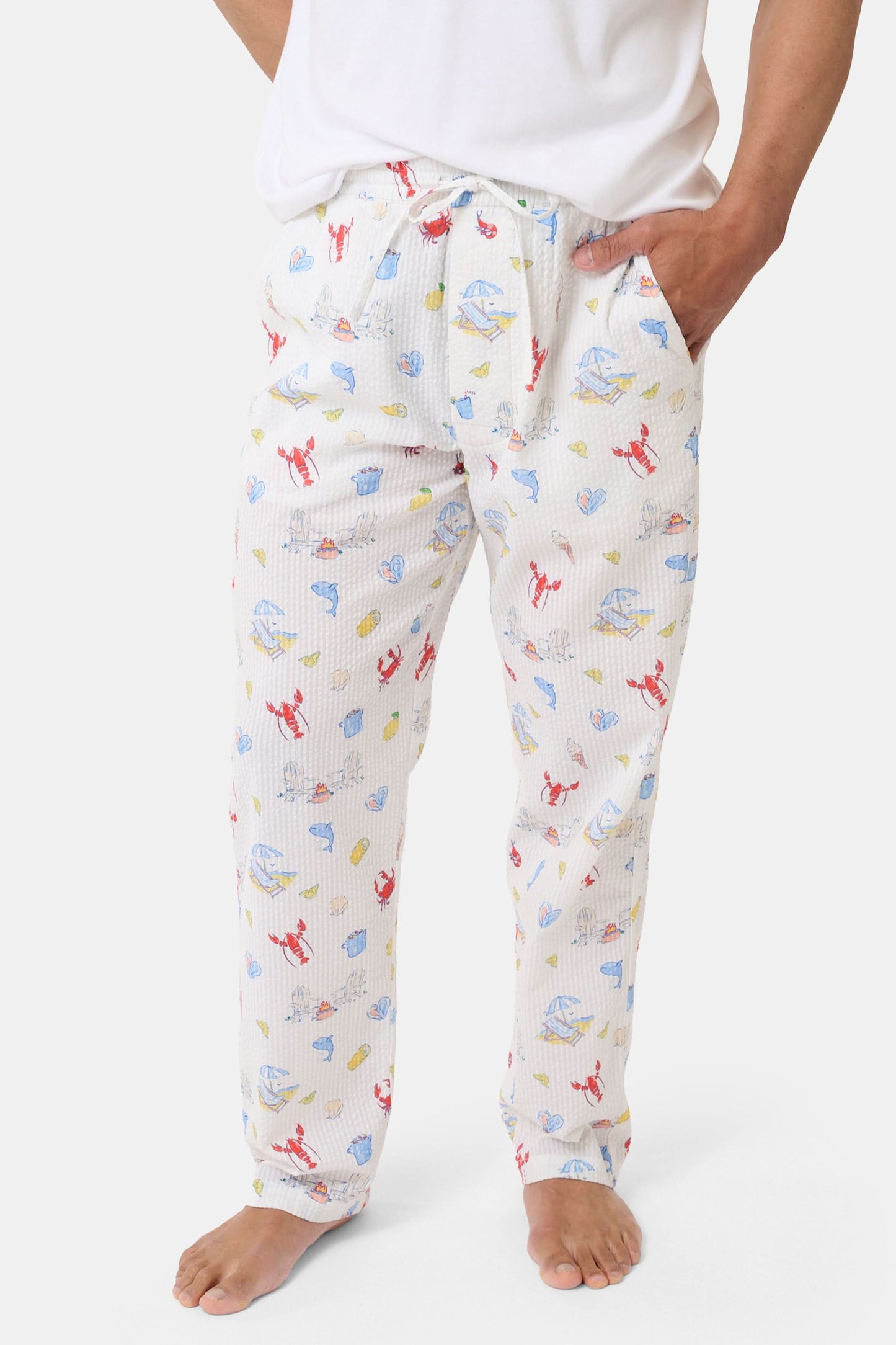 A person stands barefoot in Roller Rabbit’s Men’s Catch of the Day Jon Pant—loose white seersucker pants with a lobsters, fish, and seashells print—and a white t-shirt, one hand tucked into the pants’ pocket.