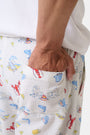 A person stands barefoot in Roller Rabbit’s Men’s Catch of the Day Jon Pant—loose white seersucker pants with a lobsters, fish, and seashells print—and a white t-shirt, one hand tucked into the pants’ pocket.