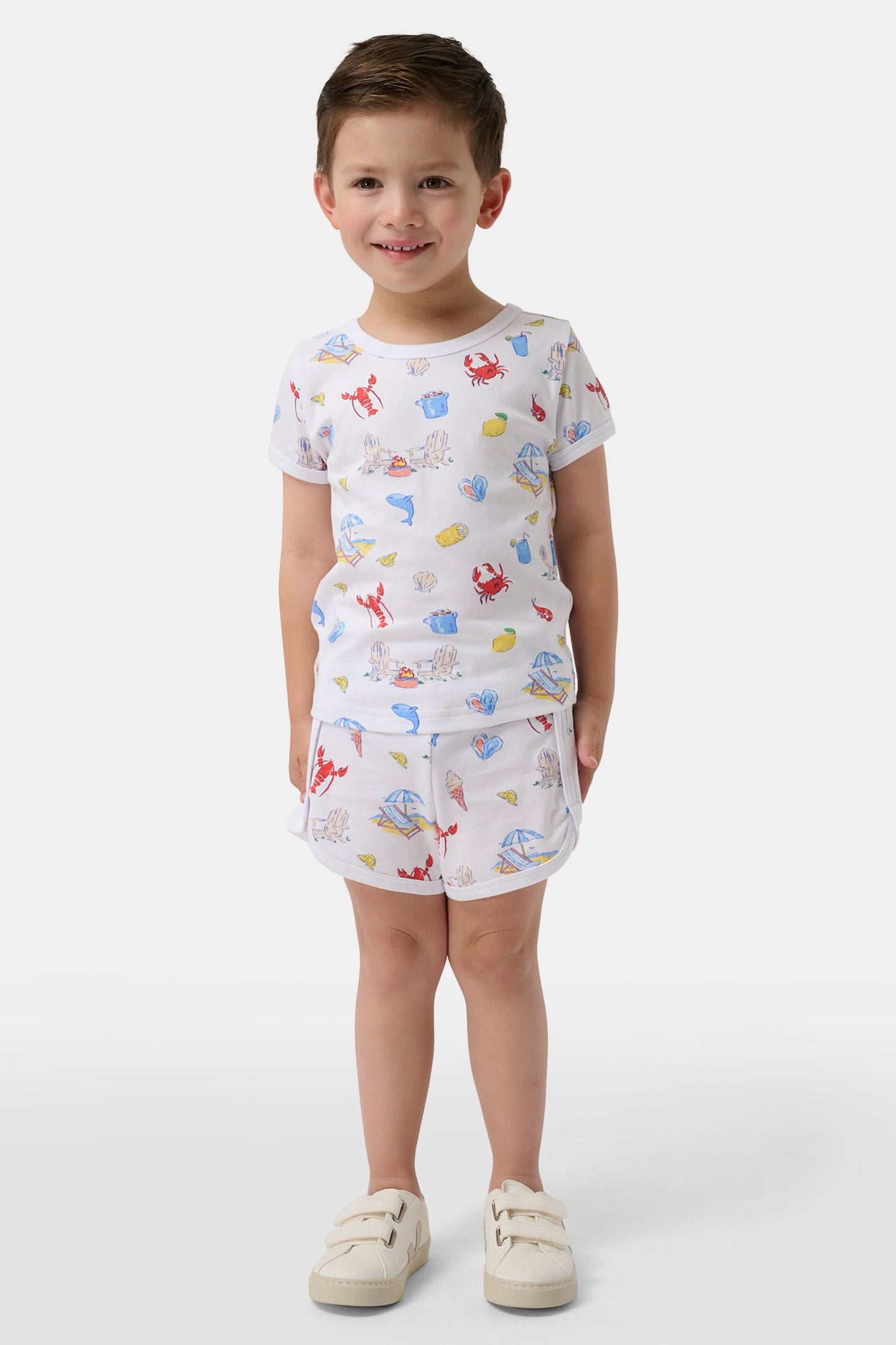 A young child smiles in a Roller Rabbit Kids Catch of the Day Playa Play Set—white t-shirt and shorts with colorful sea creature prints, paired with cream-colored shoes, against a plain white background.