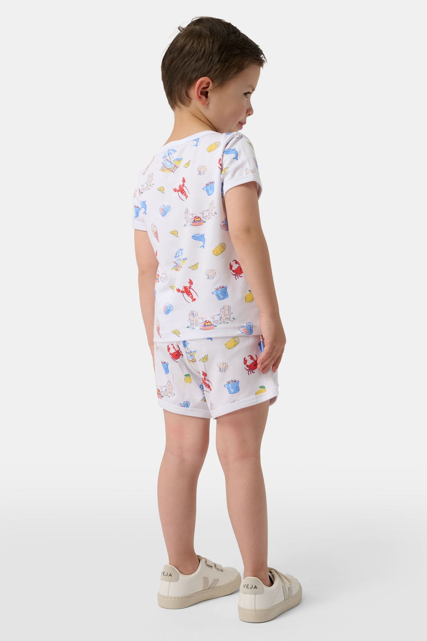 A young child smiles in a Roller Rabbit Kids Catch of the Day Playa Play Set—white t-shirt and shorts with colorful sea creature prints, paired with cream-colored shoes, against a plain white background.