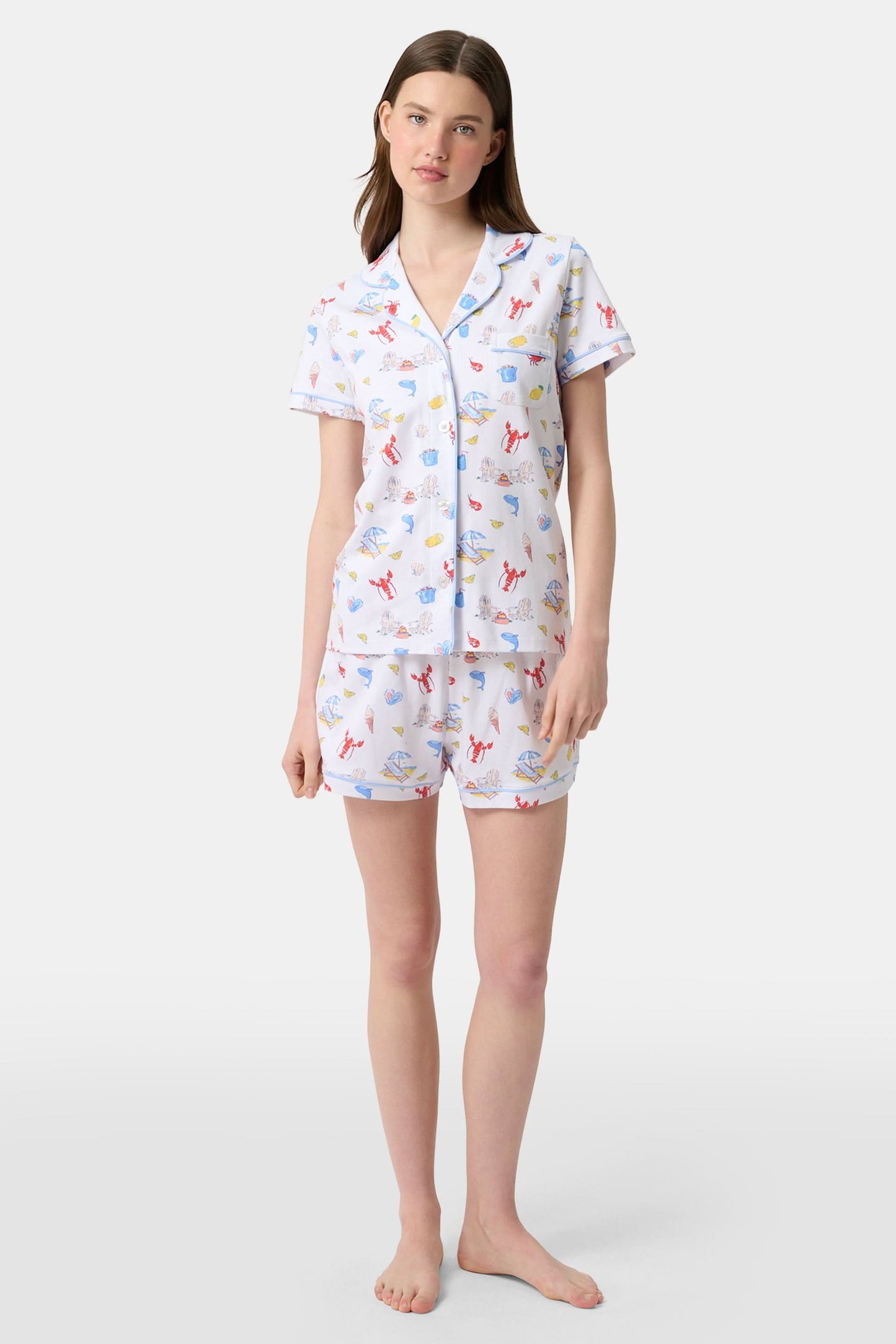 A woman stands barefoot in Roller Rabbit's Catch of the Day Polo Pajamas—short-sleeve, button-up PJs with matching shorts, both featuring a colorful seaside print—facing forward against a plain white background.
