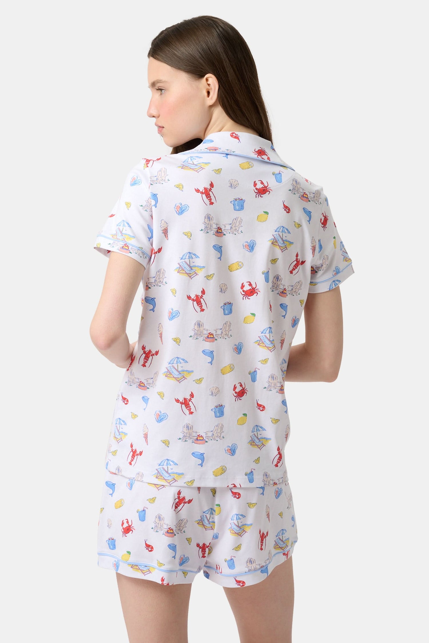 A woman stands barefoot in Roller Rabbit's Catch of the Day Polo Pajamas—short-sleeve, button-up PJs with matching shorts, both featuring a colorful seaside print—facing forward against a plain white background.