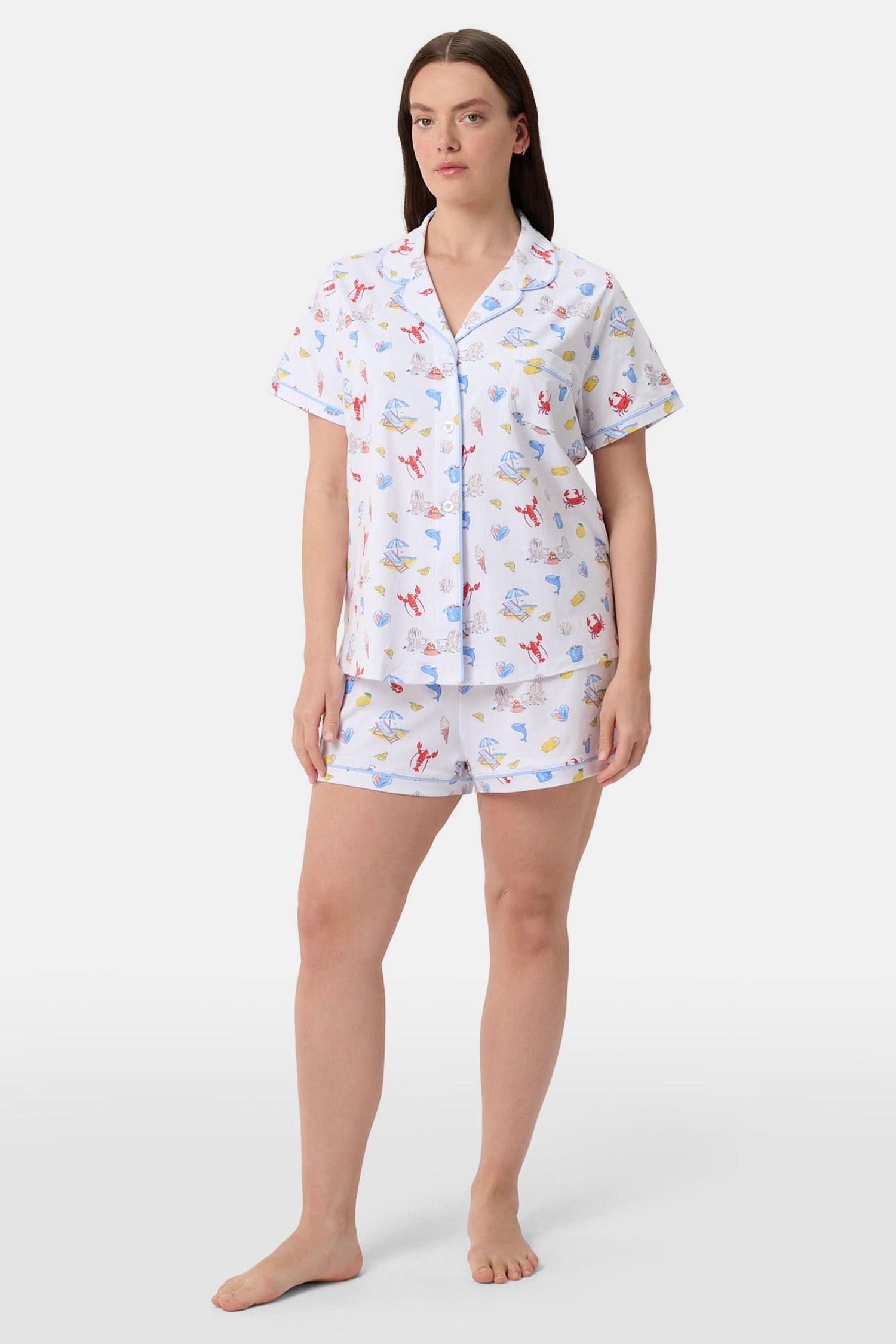 A woman stands barefoot in Roller Rabbit's Catch of the Day Polo Pajamas—short-sleeve, button-up PJs with matching shorts, both featuring a colorful seaside print—facing forward against a plain white background.