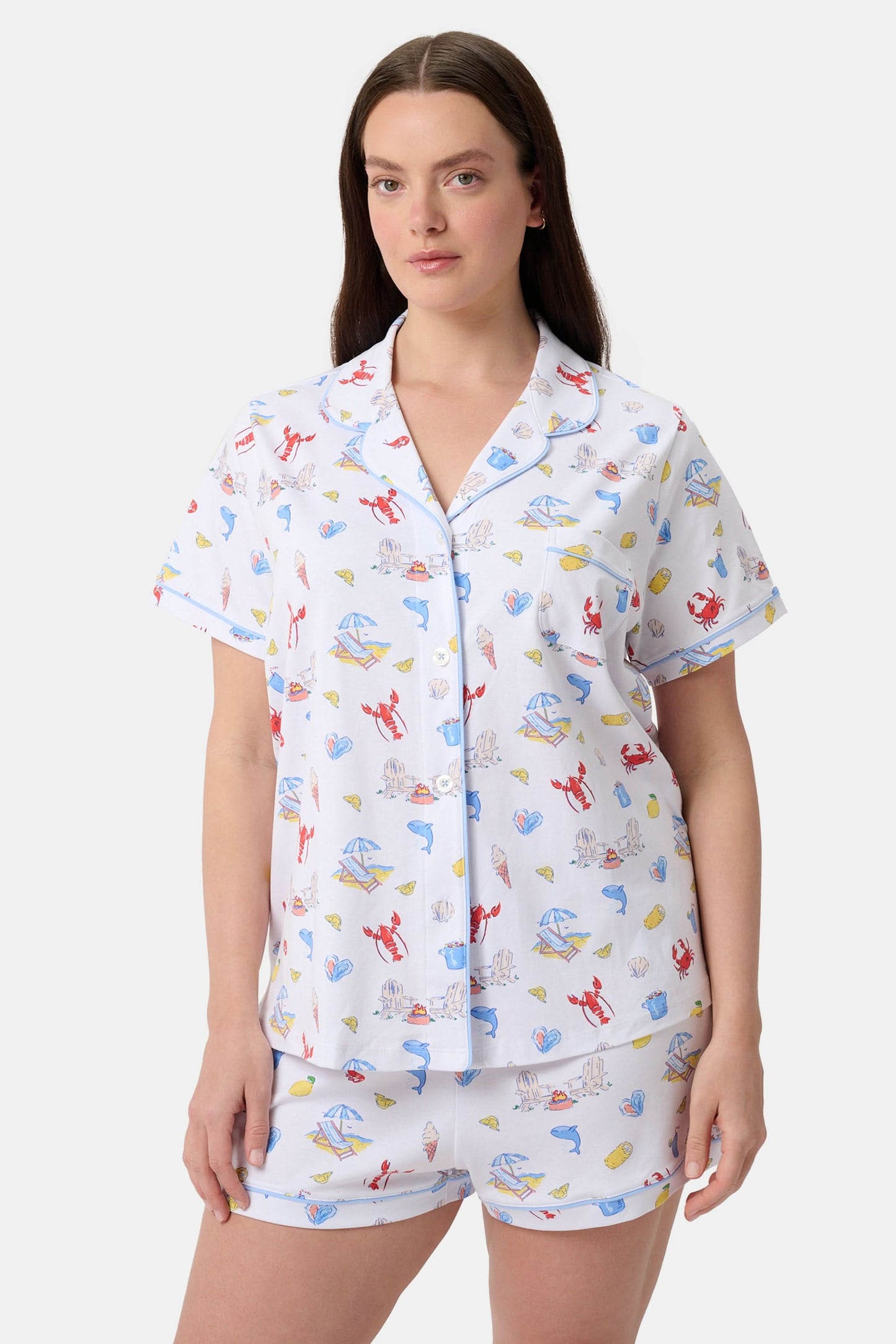 A woman stands barefoot in Roller Rabbit's Catch of the Day Polo Pajamas—short-sleeve, button-up PJs with matching shorts, both featuring a colorful seaside print—facing forward against a plain white background.