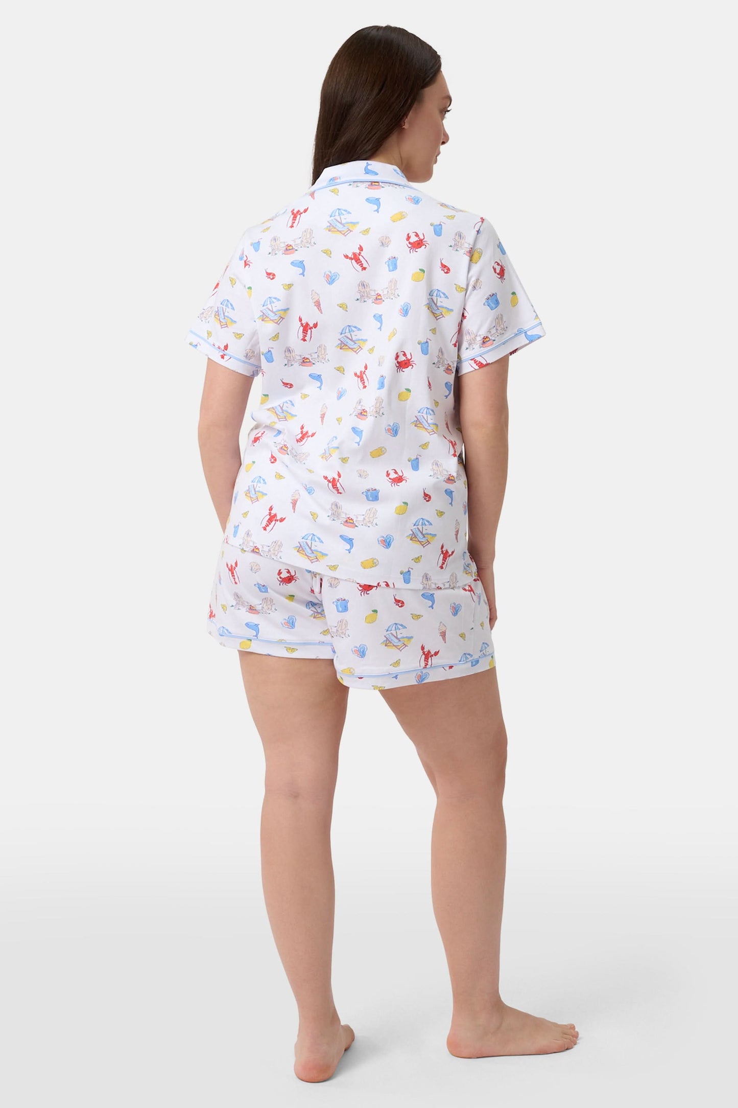 A woman stands barefoot in Roller Rabbit's Catch of the Day Polo Pajamas—short-sleeve, button-up PJs with matching shorts, both featuring a colorful seaside print—facing forward against a plain white background.