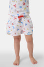 A young boy smiles barefoot in front of a cream-colored vehicle, wearing Roller Rabbit Boys Catch of the Day Shortie Swim Trunk with colorful prints, while another child sits partially visible beside him.
