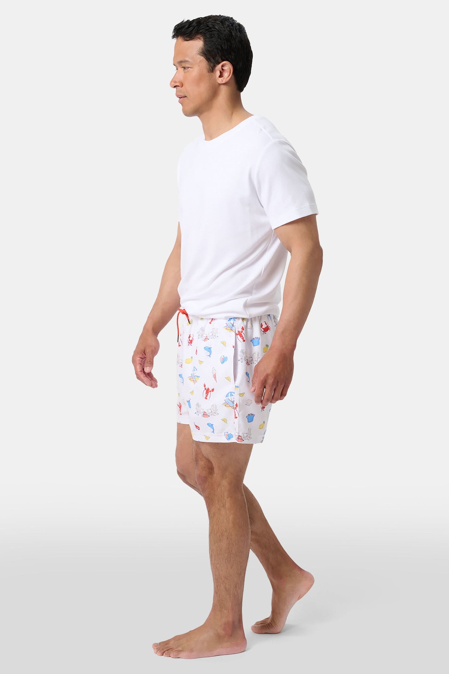 A person stands barefoot, one hand in a pocket, wearing a white T-shirt and Roller Rabbit Men's Catch of the Day Swim Trunk with colorful beach-themed prints, an elastic waistband, and a classic fit.