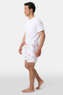 A person stands barefoot, one hand in a pocket, wearing a white T-shirt and Roller Rabbit Men's Catch of the Day Swim Trunk with colorful beach-themed prints, an elastic waistband, and a classic fit.