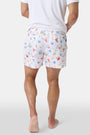 A person stands barefoot, one hand in a pocket, wearing a white T-shirt and Roller Rabbit Men's Catch of the Day Swim Trunk with colorful beach-themed prints, an elastic waistband, and a classic fit.