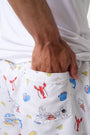 A person stands barefoot, one hand in a pocket, wearing a white T-shirt and Roller Rabbit Men's Catch of the Day Swim Trunk with colorful beach-themed prints, an elastic waistband, and a classic fit.