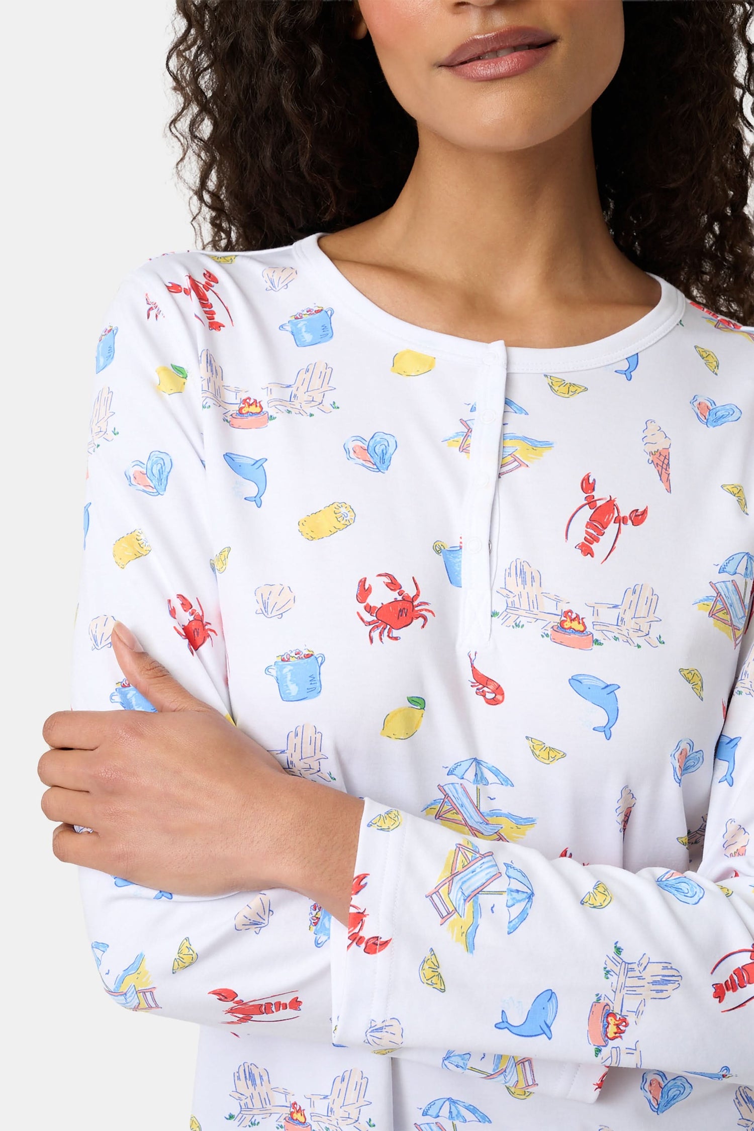A woman with curly hair wears Roller Rabbit Catch of the Day Pajamas, made from soft Pima cotton and featuring colorful cartoon animal prints. She stands barefoot on a plain white background, facing forward with a neutral expression.