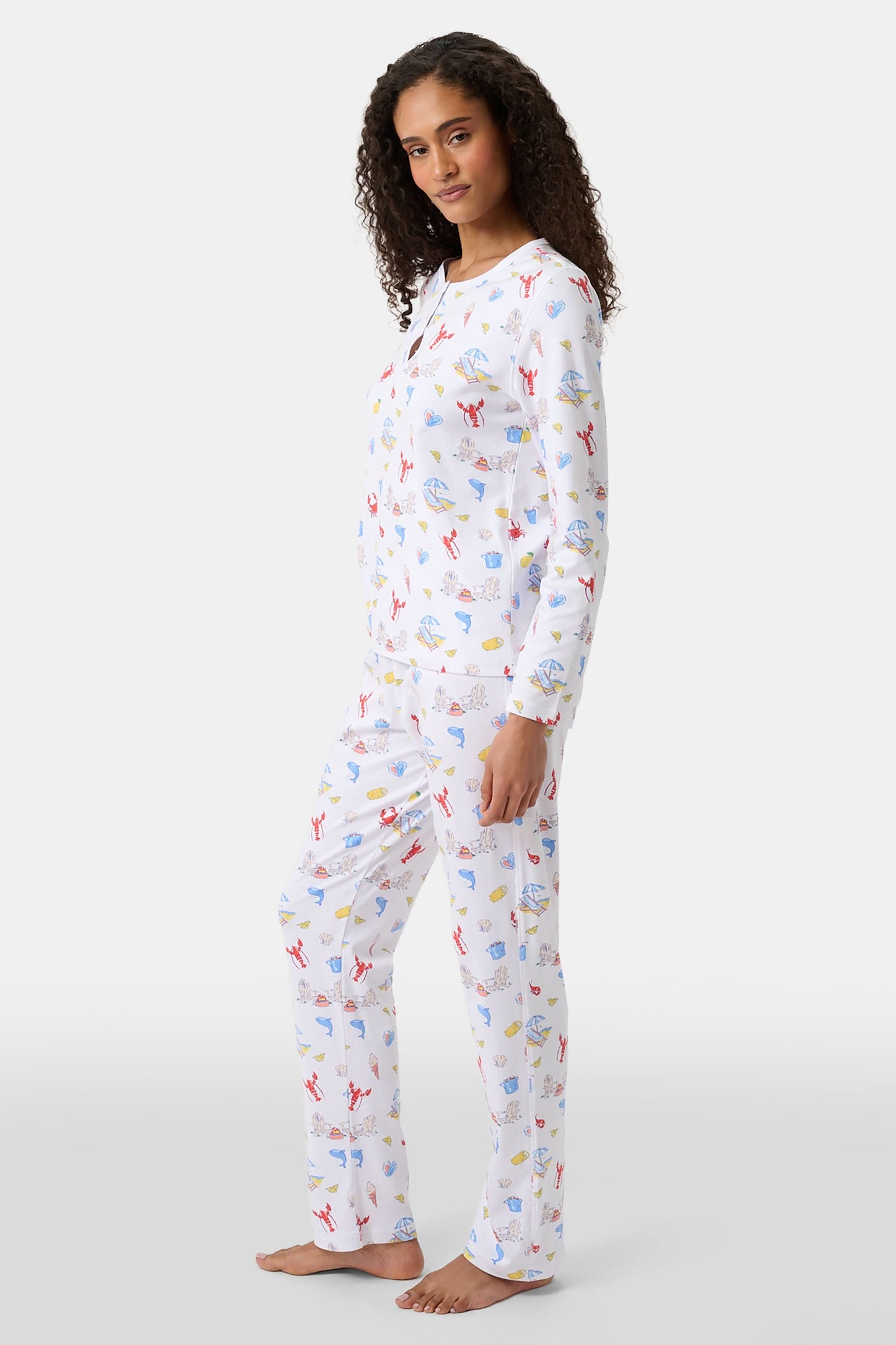 A woman with curly hair wears Roller Rabbit Catch of the Day Pajamas, made from soft Pima cotton and featuring colorful cartoon animal prints. She stands barefoot on a plain white background, facing forward with a neutral expression.