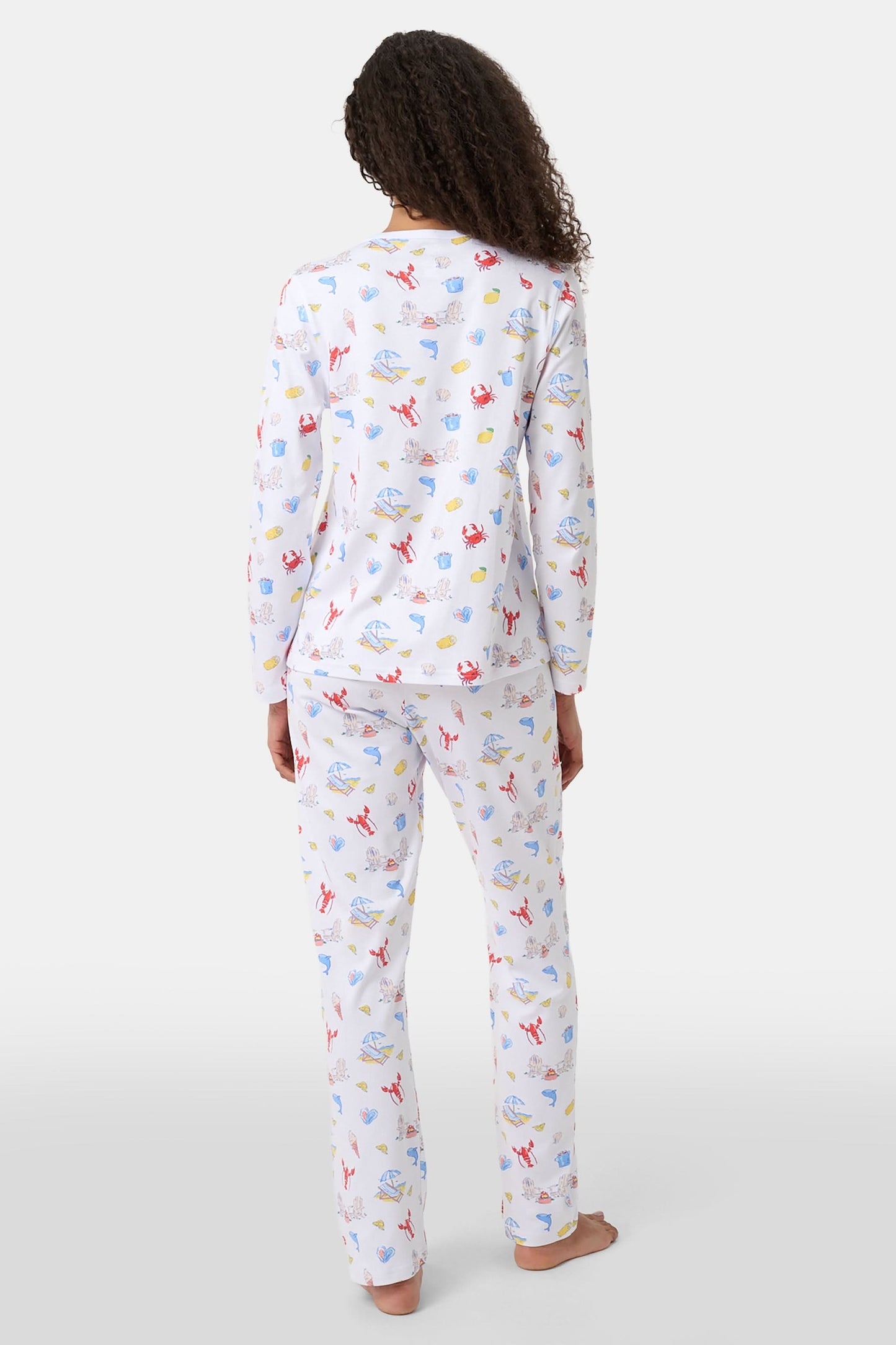A woman with curly hair wears Roller Rabbit Catch of the Day Pajamas, made from soft Pima cotton and featuring colorful cartoon animal prints. She stands barefoot on a plain white background, facing forward with a neutral expression.