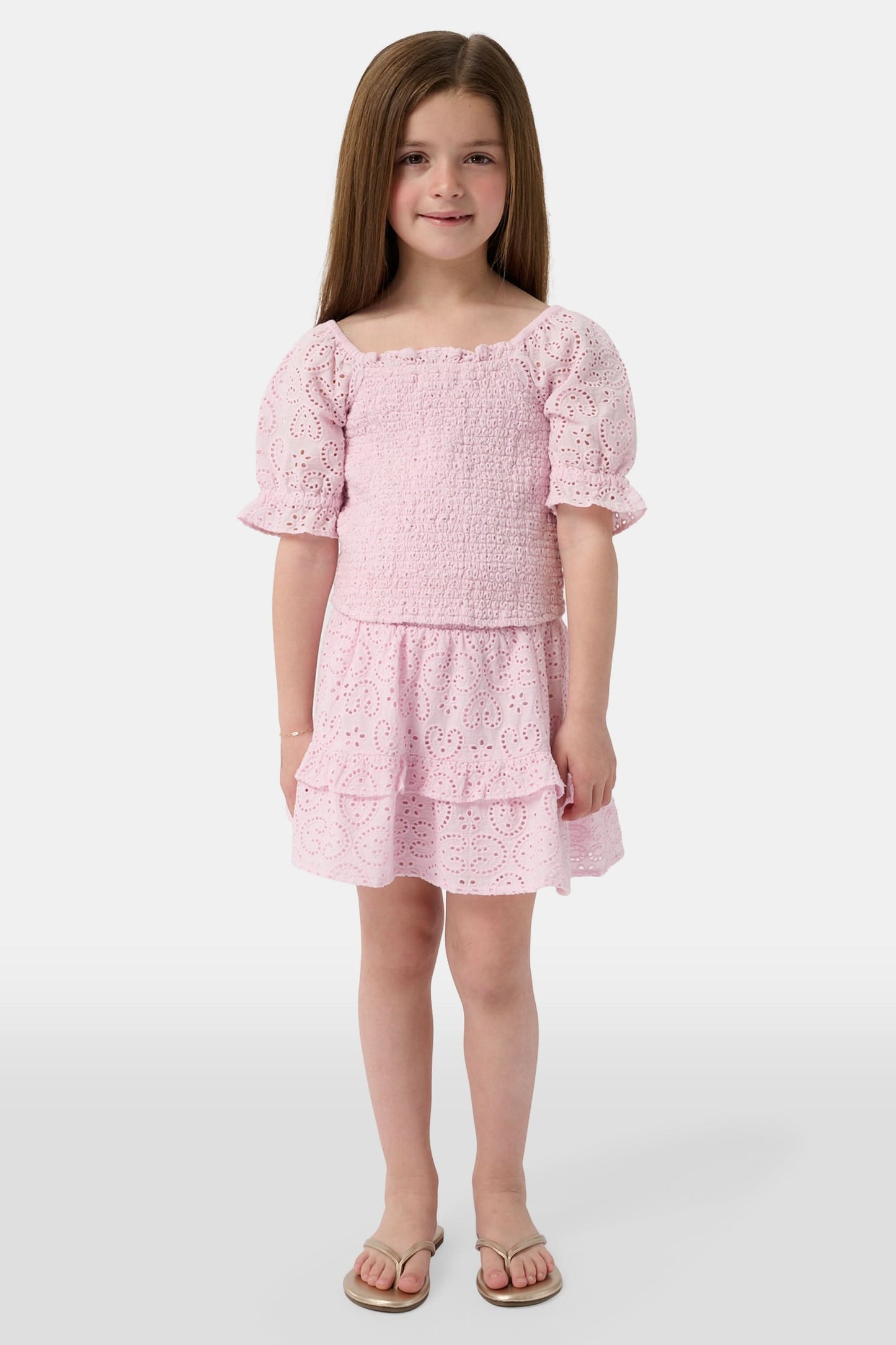 A young girl with long brown hair is wearing the Roller Rabbit Kids Clover Eyelet Elsie Top, a light pink, short-sleeved, smocked kids top, paired with a matching heart-patterned eyelet skirt. She smiles against a plain light background.
