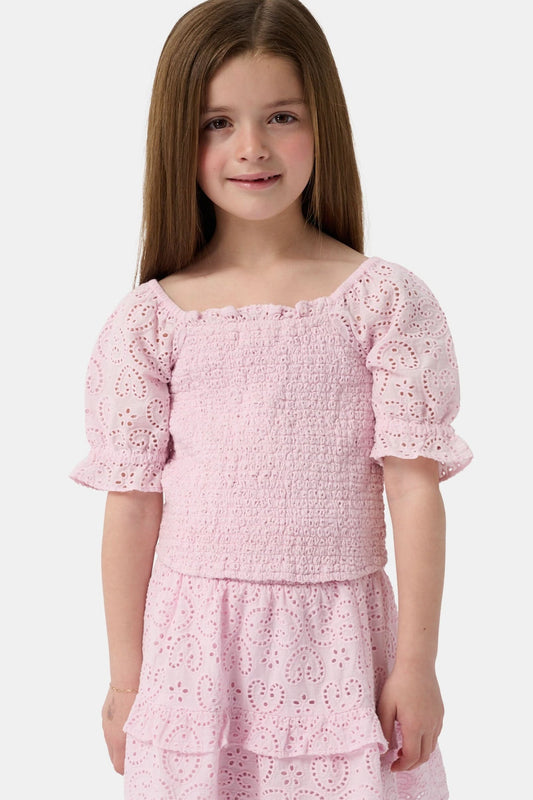 A young girl with long brown hair is wearing the Roller Rabbit Kids Clover Eyelet Elsie Top, a light pink, short-sleeved, smocked kids top, paired with a matching heart-patterned eyelet skirt. She smiles against a plain light background.