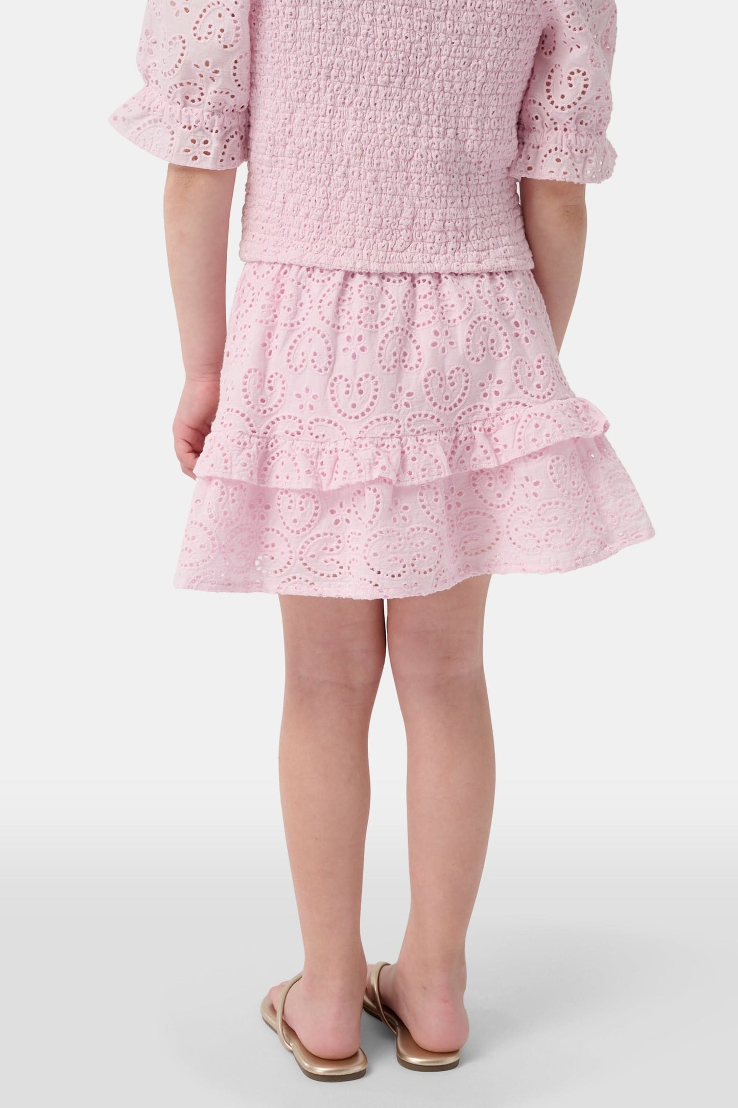 A young child wears the Roller Rabbit Kids Clover Eyelet Emry Skirt in light pink with ruffles and a matching smocked top, standing with their left arm slightly bent against a plain white background.