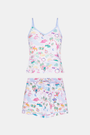 A woman with long brown hair wears the Roller Rabbit Camp RR Mischa Set, a light-hued Pima cotton pajama set with playful prints. The adjustable strap tank and shorts provide comfort as she stands turned slightly to the side, gazing off-camera.