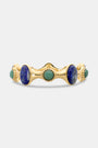 The Bejeweled Bangle by Roller Rabbit features oversized oval blue and green semi-precious stones set in a wavy, textured gold band with beaded details, shown against a plain white background.