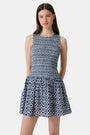 A woman with long straight hair and a neutral expression wears the Roller Rabbit Diamond Ikat Isla Dress—a navy smocked sleeveless dress with a white ikat pattern, flared skirt, and dropped waist, standing against a plain background.