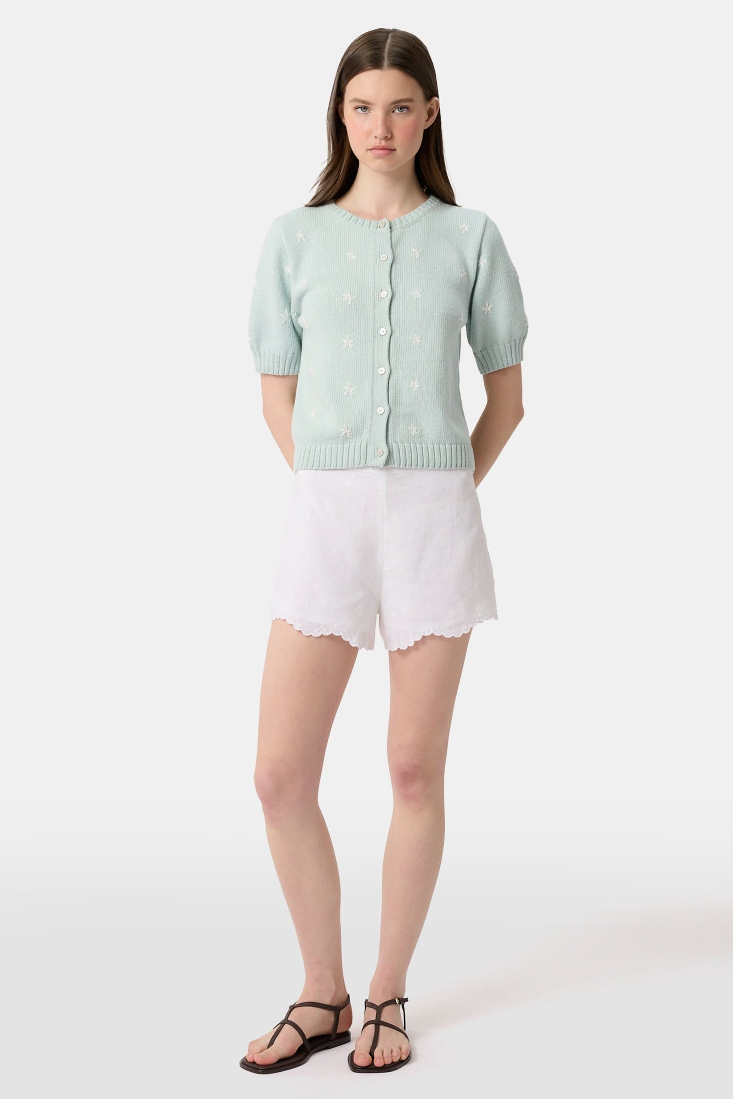 A person wears the Roller Rabbit Ditsy Floral Embroidery Jordana Shorts—white, high-waisted, with a subtle floral design and scalloped lace trim—paired with a light green buttoned sleeveless knit top. The background is plain white.