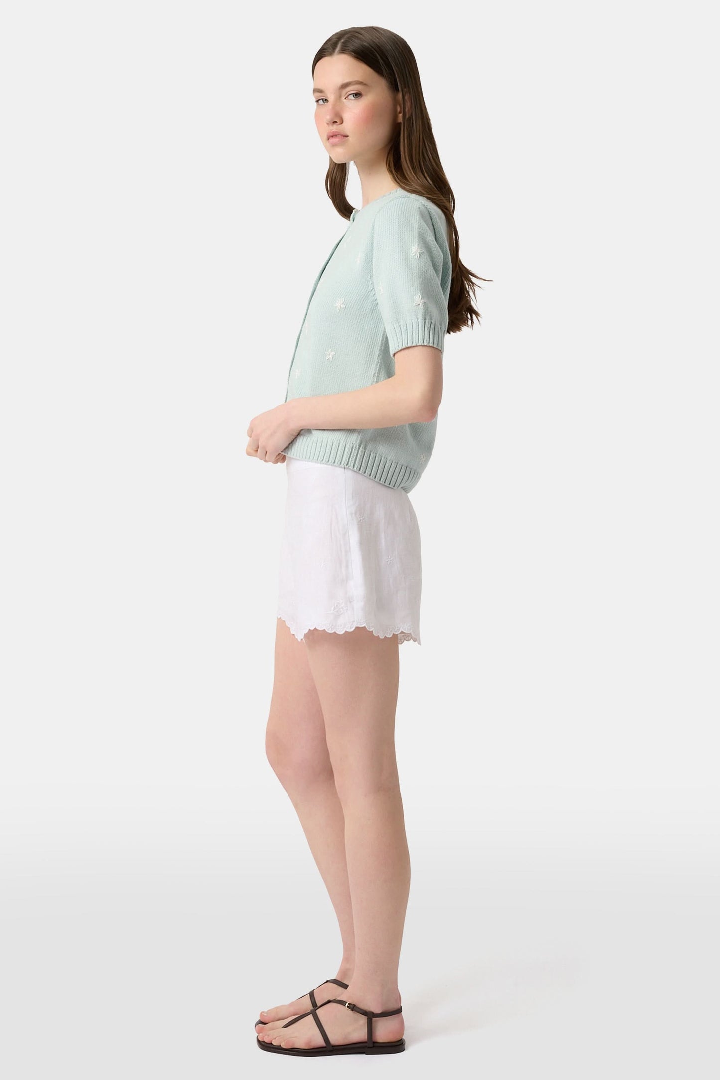 A person wears the Roller Rabbit Ditsy Floral Embroidery Jordana Shorts—white, high-waisted, with a subtle floral design and scalloped lace trim—paired with a light green buttoned sleeveless knit top. The background is plain white.