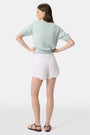 A person wears the Roller Rabbit Ditsy Floral Embroidery Jordana Shorts—white, high-waisted, with a subtle floral design and scalloped lace trim—paired with a light green buttoned sleeveless knit top. The background is plain white.