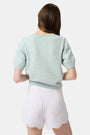 A person wears the Roller Rabbit Ditsy Floral Embroidery Jordana Shorts—white, high-waisted, with a subtle floral design and scalloped lace trim—paired with a light green buttoned sleeveless knit top. The background is plain white.