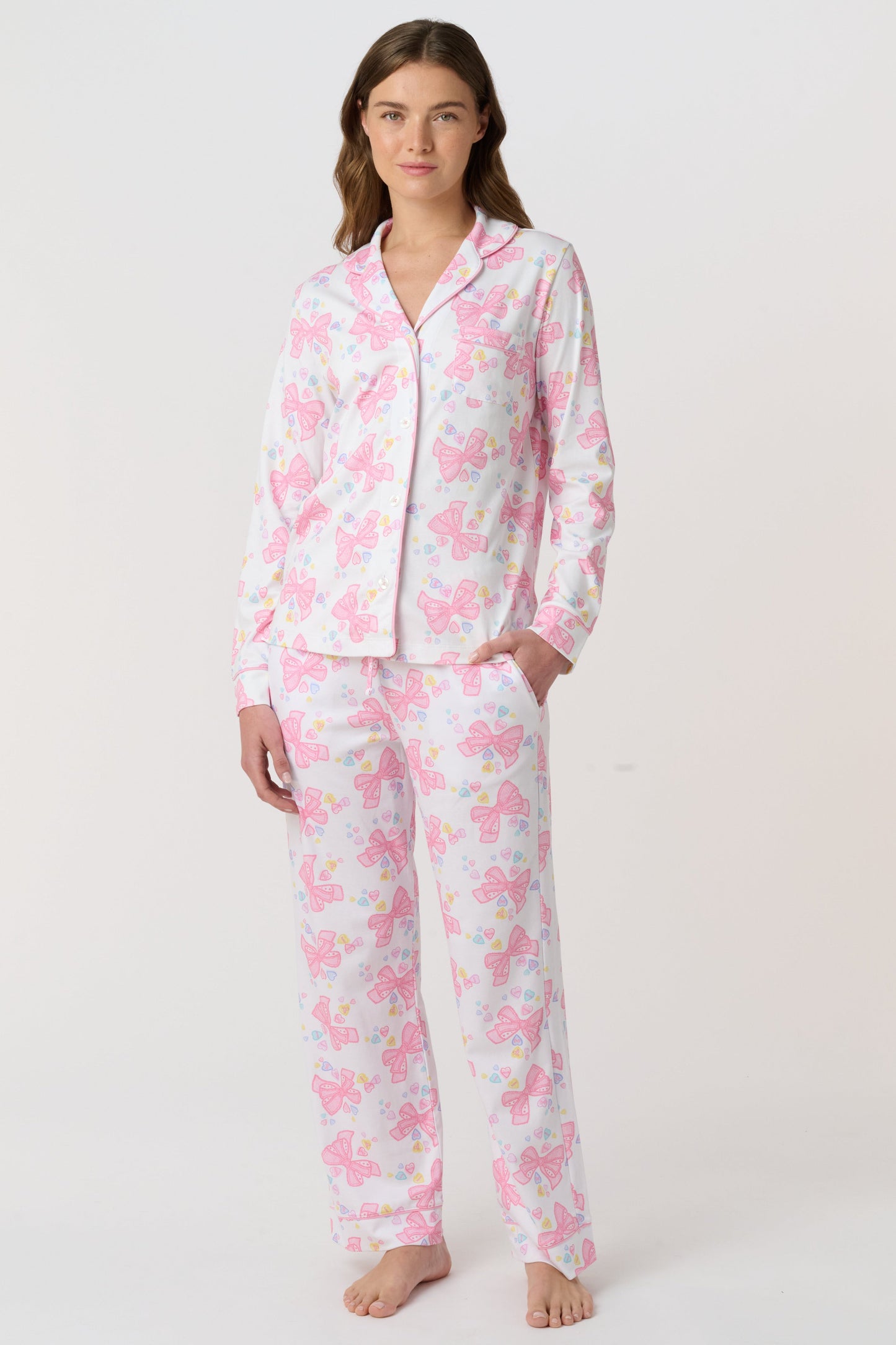 A woman stands barefoot, facing the camera in Roller Rabbit Doily Beaux Long Sleeve Polo Pajamas featuring a pink print. The matching set includes a button-up top and long pants; she wears a neutral expression.