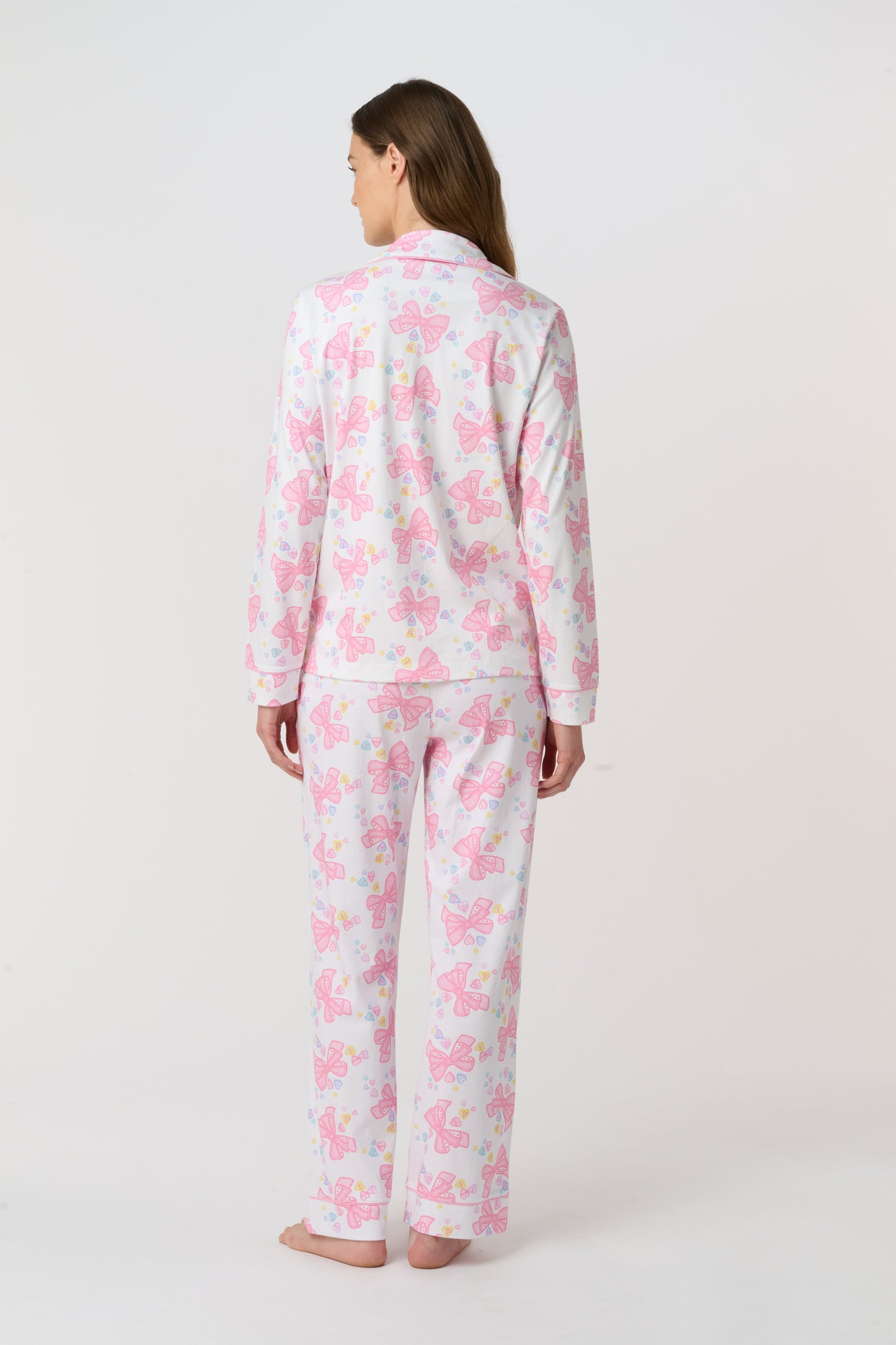 A woman stands barefoot, facing the camera in Roller Rabbit Doily Beaux Long Sleeve Polo Pajamas featuring a pink print. The matching set includes a button-up top and long pants; she wears a neutral expression.