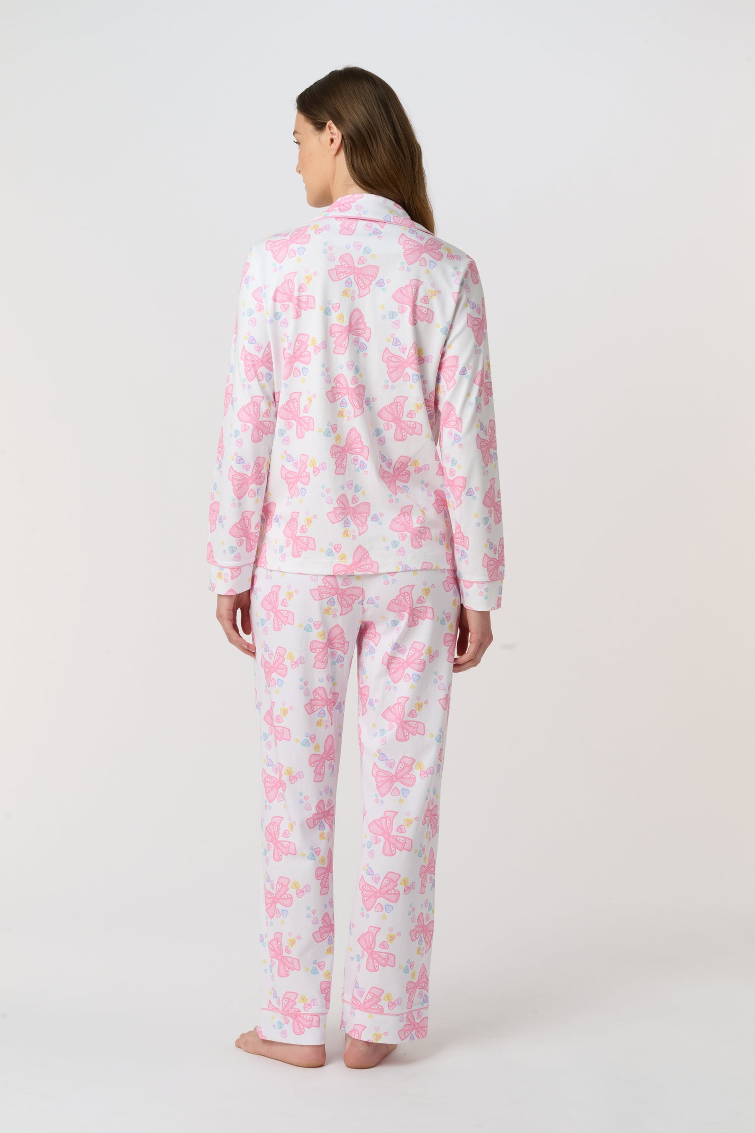 A woman stands barefoot, facing the camera in Roller Rabbit Doily Beaux Long Sleeve Polo Pajamas featuring a pink print. The matching set includes a button-up top and long pants; she wears a neutral expression.