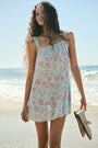 A woman with curly hair wears the Camellia Block Keaton Dress by Roller Rabbit, a sleeveless, loose-fit 100 cotton sundress with pink floral print on a light green background. She smiles softly with her hands behind her back.