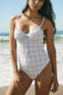 A woman wearing the Roller Rabbit Sun Check Eyelet Aurora One Piece, a light blue and white plaid swimsuit with adjustable straps, stands on a beach with waves and a blue sky in the background.