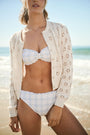 A woman with long dark hair smiles in the Roller Rabbit Floral Crochet Moxie Cardigan, featuring flower-like holes; she pairs it with light floral ruffled-hem shorts and rests one hand in her pocket.