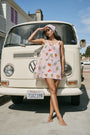 A woman in the Roller Rabbit Fruits D'Ete Keaton Dress with a matching headscarf stands barefoot by a vintage white Volkswagen van, shielding her eyes from the sun. The sunny coastal backdrop highlights the dress's playful print.
