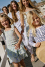 A girl with long curly hair wears the Kids Zelda Floral Tory Top by Roller Rabbit, styled as a light blue and green tiered dress, standing against a plain white background with her hands behind her back.