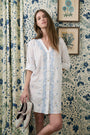 A woman wearing the Roller Rabbit Vanna Embroidery Janna Cover Up stands against a floral-patterned wall and curtain, holding shoes in her left hand. Two framed botanical prints hang on the wall behind her.