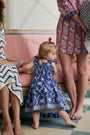 A young girl with straight brown hair smiles while wearing the Roller Rabbit Kids Lobster Trellis Ceri Dress, featuring a blue and white pattern with ruffled sleeves, against a plain white background.
