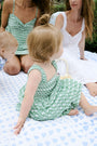 A young girl with curly hair wears the Roller Rabbit Kids Cane Kenia Dress, a green and white patterned cotton tiered dress with wide straps, standing against a plain light background.