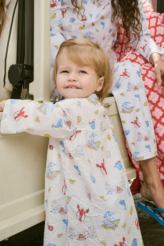 A young child with light brown hair smiles near an open car door, wearing the Roller Rabbit Kids Catch of the Day Serafina Tunic. An adult in matching pajamas is partially visible behind them.