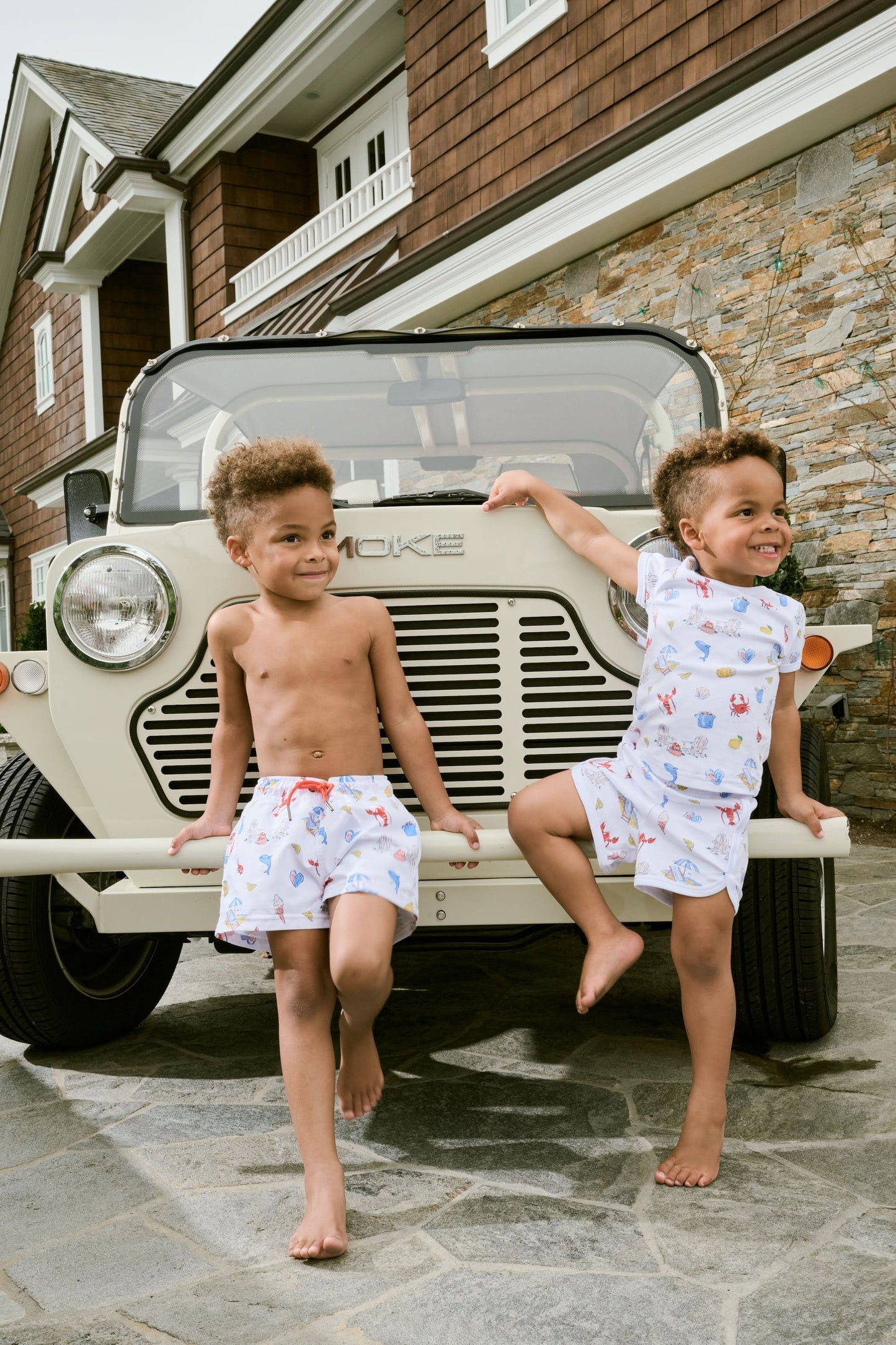 A young child smiles in a Roller Rabbit Kids Catch of the Day Playa Play Set—white t-shirt and shorts with colorful sea creature prints, paired with cream-colored shoes, against a plain white background.