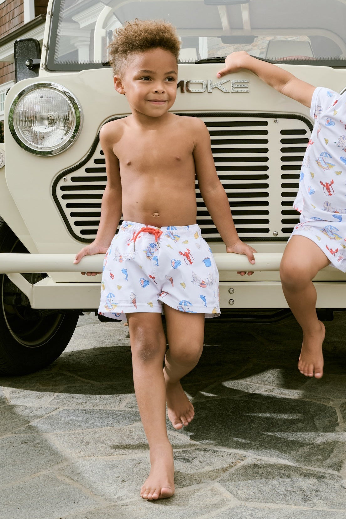 A young boy smiles barefoot in front of a cream-colored vehicle, wearing Roller Rabbit Boys Catch of the Day Shortie Swim Trunk with colorful prints, while another child sits partially visible beside him.