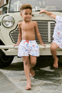 A young boy smiles barefoot in front of a cream-colored vehicle, wearing Roller Rabbit Boys Catch of the Day Shortie Swim Trunk with colorful prints, while another child sits partially visible beside him.