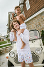A person stands barefoot, one hand in a pocket, wearing a white T-shirt and Roller Rabbit Men's Catch of the Day Swim Trunk with colorful beach-themed prints, an elastic waistband, and a classic fit.